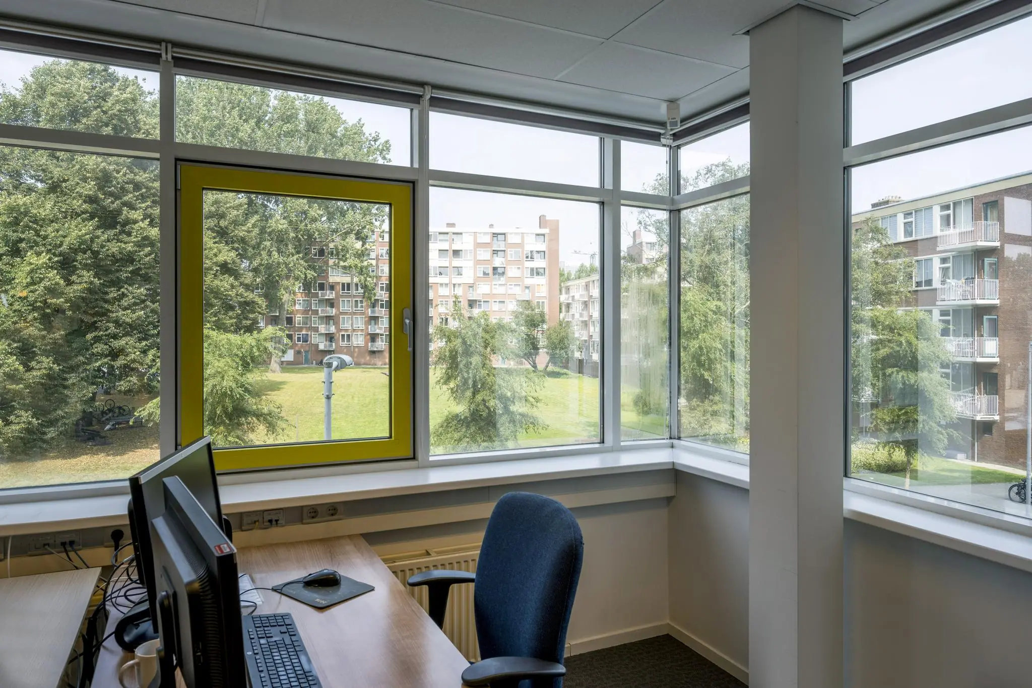 Kantoorruimte met werkplek en computers, met uitzicht op een groen binnenterrein en flatgebouwen aan de Jan Tooropstraat.